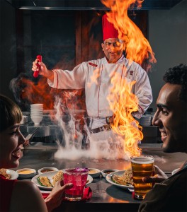 Benihana chef creates dramatic flame at the grill while a couple in the foreground talk and enjoy drinks at the teppanyaki table.